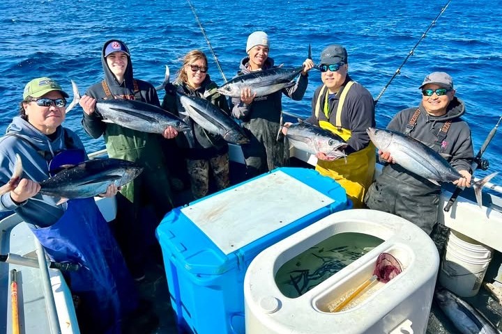 Group of six people on a boat holding freshly caught fish over the ocean.