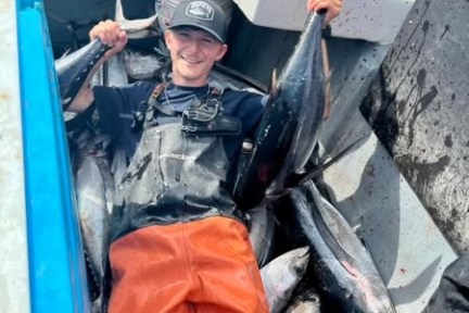 Person in orange overalls holding two fish, sitting among several fish on a boat.