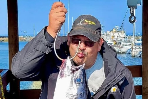 Man kissing a large fish hooked on a scale, wearing a hat and sunglasses, with a marina in the background.