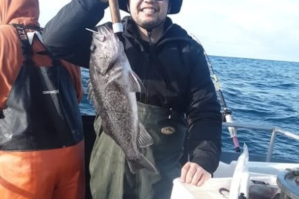 Person in hat and sunglasses holds a large fish on a boat with another person nearby, ocean in background.