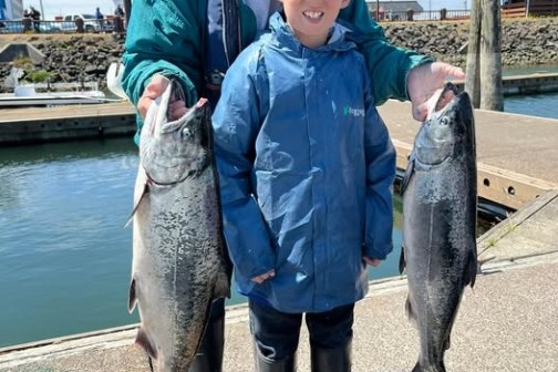 Person and child holding two large fish on a dock near the water.
