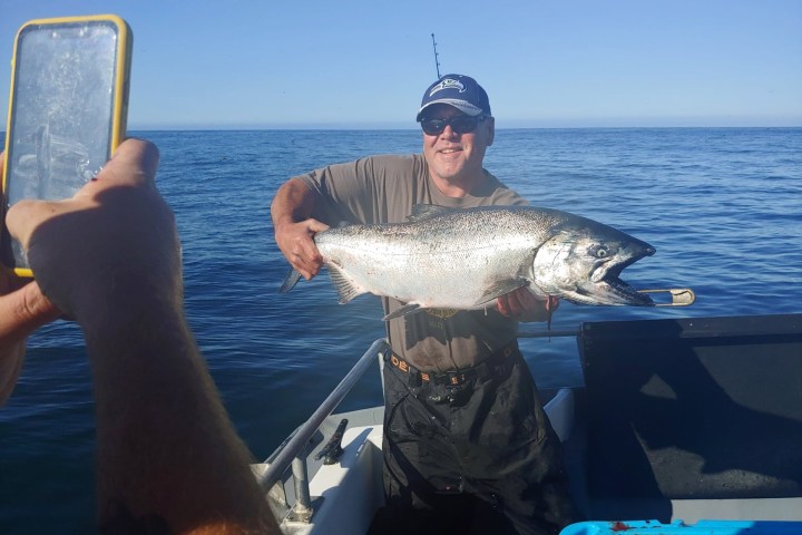 Man on boat holding large fish, another person taking a photo with a smartphone.