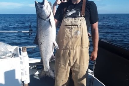 Person in overalls on a boat holding a large fish, smiling against a backdrop of ocean and blue sky.