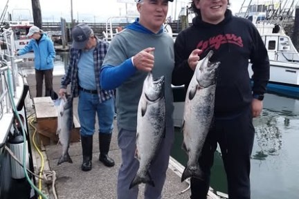 Two people holding fish on a dock, with boats and additional people in the background.