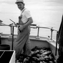 Person in overalls on a boat with fish on the deck, holding a tool, overcast sky in the background.