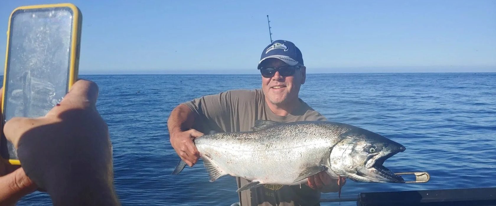 Man holding a large fish on a boat, while someone takes a photo with a smartphone.