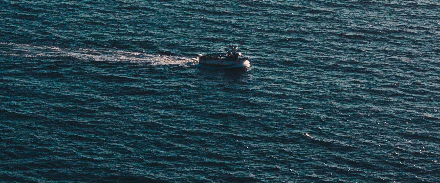 A small boat travels across a vast, calm blue ocean.
